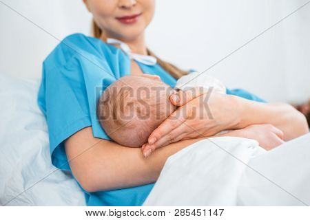 Low Angle View Of Smiling Young Mother Breastfeeding Newborn Baby On Bed In Hospital Room