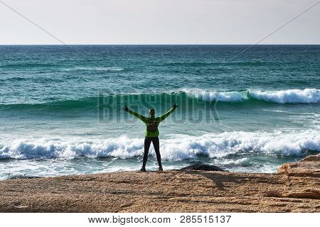 A Male Athlete With Arms Outstretched To The Side Is Standing On The Seashore. View From The Back. J