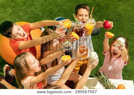 Kids Fashion Concept. The Group Of Teen Boys And Girls Sitting On Green Grass At Park. Children Colo