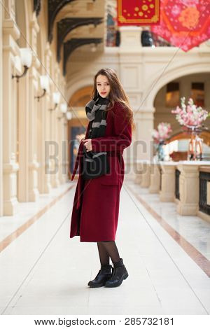 Full body portrait. Young beautiful woman in stylish in a long burgundy coat, indoor shop