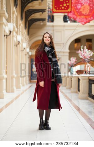 Full body portrait. Young beautiful woman in stylish in a long burgundy coat, indoor shop