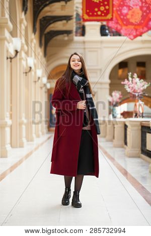 Full body portrait. Young beautiful woman in stylish in a long burgundy coat, indoor shop