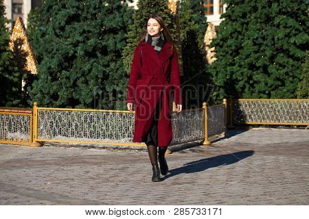 Full body portrait. Young beautiful brunette woman in stylish in a long burgundy coat walking in spring street