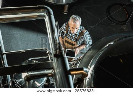 Overhead View Of Professional Senior Brewer Using Digital Tablet And Working With Brewery Equipment