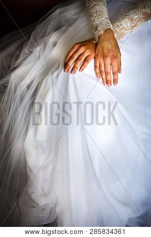 Crossed Feamle Hands Are Laying On The White Wedding Dress Closeup.