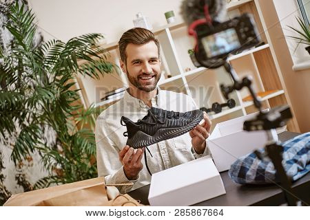 Sport Shoes. Cheerful Male Blogger Holding Black Sneakers While Recording New Video For His Blog.