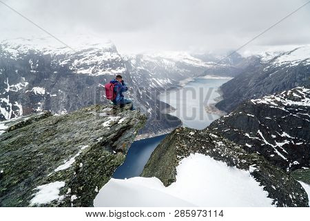 Trolltunga Cliff Under Snow In Norway. Scenic Landscape. Man Traveller Sitting On Edge Of Rock And M