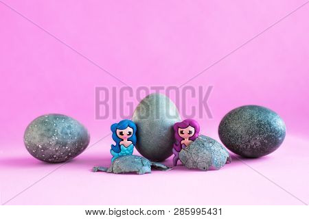 Three Gray, Blue Easter Eggs, Like Stones. Two Toy Mermaids. Lilac Background