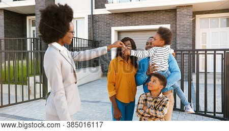 Female Real-estate Selling House To African American Happy Family With Kids. Handing Keys. Outdoors.