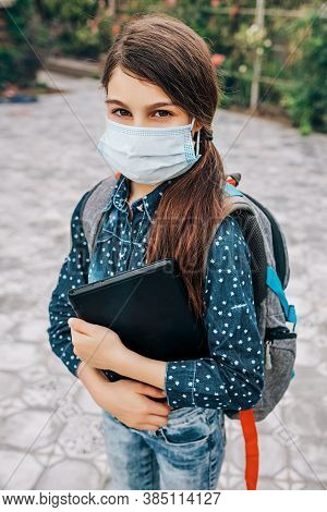 Little Schoolgirl With A Medical Mask On Her Face And A Laptop In Her Hand Goes To School During The