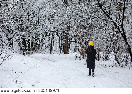 Woman In Black Winter Coat And Yellow Hat Walking By Snowed City Park
