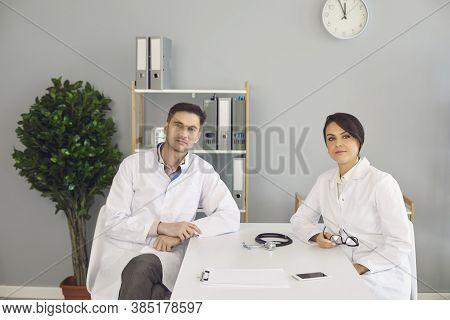 Woman And Man Doctors Sitting And Looking At Camera In Medical Clinic