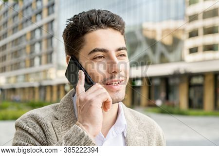 Happy young mixed-race businessman in formalwear talking to clients on mobile phone in urband environment against modern architecture