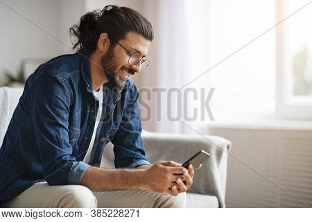 New Mobile App. Stylish Western Millennial Guy Using Smatphone While Sitting On Sofa At Home, Browsi