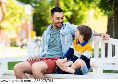 family, fatherhood and people concept - happy father and son sitting on bench at summer park and talking