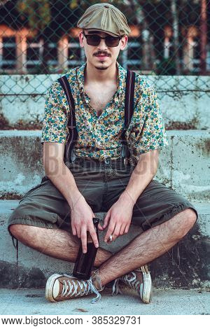 Young Hipster Man In Retro Clothing Sits On The Stairs With Beer In His Hands