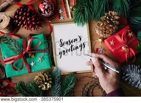 Human Hand Writing Season's Greeting Text On Cards With Gift Box Present And Ornament Element On Woo