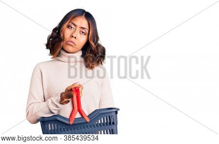 Young beautiful mixed race woman holding supermarket shopping basket thinking attitude and sober expression looking self confident 