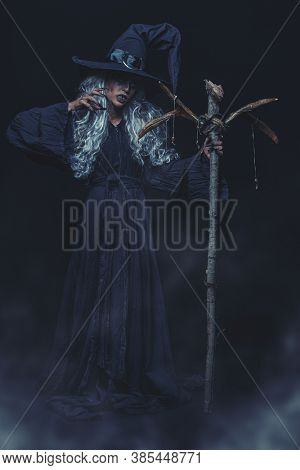 Halloween. Full length portrait of an ugly old witch standing with her magic staff on a black backgroung. Mysterious fog around. Witchcraft.