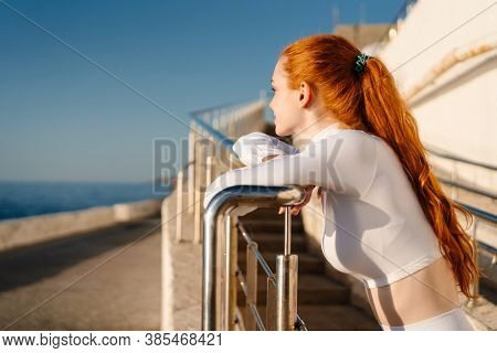 Image of redhead young sportswoman looking forward and leaning on railing at sunrise outdoors