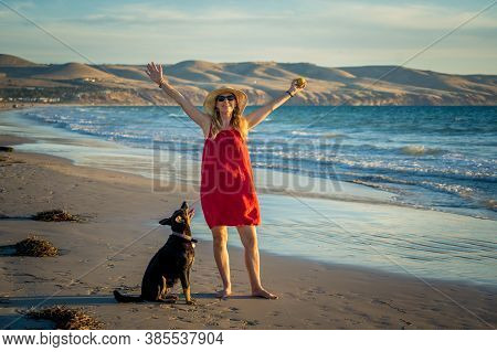 Happy Attractive Mature Woman With Her Pet Walking On Friendly Dog Beach At Sunset