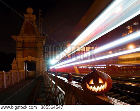 Halloween Pumpkin With A Glowing Grimace At Night On The Railway Bridge. In The Background There Are
