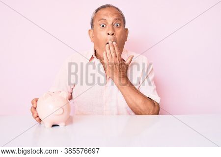 Senior handsome man with gray hair holding piggy bank sitting on the table covering mouth with hand, shocked and afraid for mistake. surprised expression 