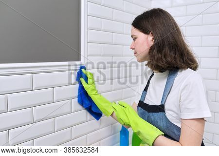 Girl Doing Cleaning In Bathroom. Teenager In Apron Gloves With Detergent And Rag Washing White Tile 