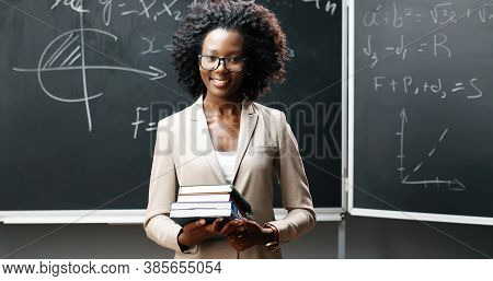 Portrait Of Young African American Female Teacher In Glasses Looking At Camera In Classrom And Holdi