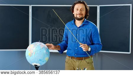 Portrait Of Caucasian Male Teacher Standing At Board With Pointer And Pointing On Globe In Class. Ma