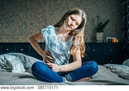 Young Brunette Girl Sits On A Bed In Her Room, Holds Her Right Side With Her Hands.