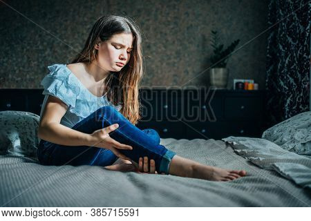 Young Girl Sits On A Bed In Her Room, Holds Her Injured Leg With Her Hand.
