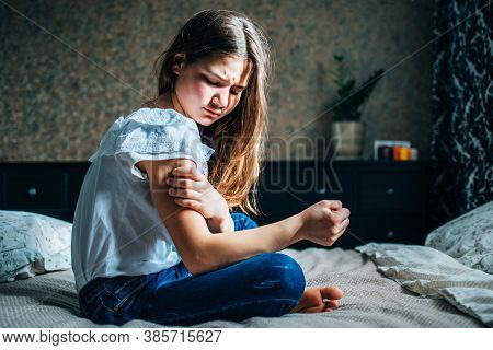 Young Brunette Girl Sitting On The Bed In Her Room, Holds An Injured Shoulder With Her Hand, Horizon