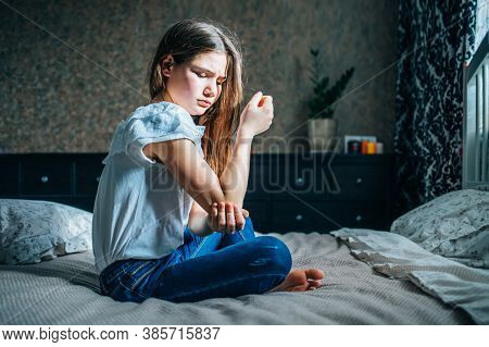 Young Brunette Girl Sits On A Bed In Her Room, Holds A Sore Elbow With Her Hand
