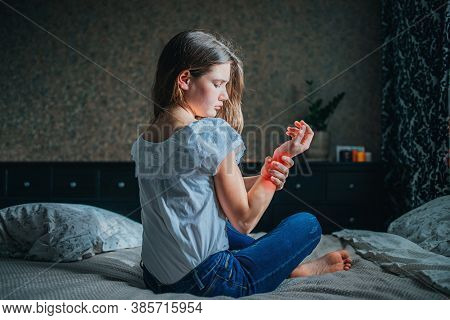 Young Dark-haired Girl Clings To Sore Wrist While Sitting Jn Bed In Her Room. Horizontal Shot, Selec