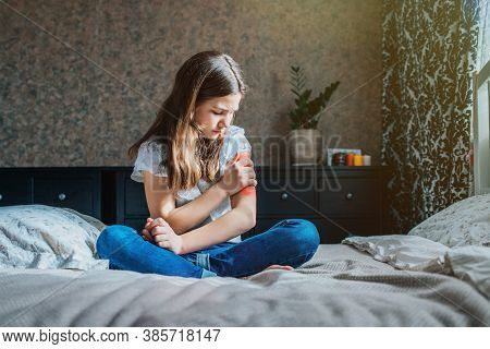 Young Brunette Girl Sits On A Bed In Her Room, Holds Her Injured Shoulder With Her Hand