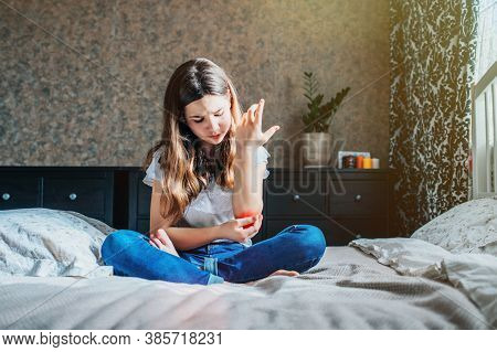 Young Brunette Girl Sits On A Bed In Her Room, Holds A Sore Elbow With Her Hand