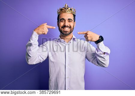 Young handsome man with beard wearing golden crown of king over purple background smiling cheerful showing and pointing with fingers teeth and mouth. Dental health concept.
