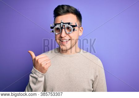 Young handsome caucasian man wearing optometrical glasses over purple background smiling with happy face looking and pointing to the side with thumb up.