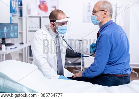 Doctor Using Stethoscope To Listen Senior Man Heart During Examination In Hospital Room And Wearing 