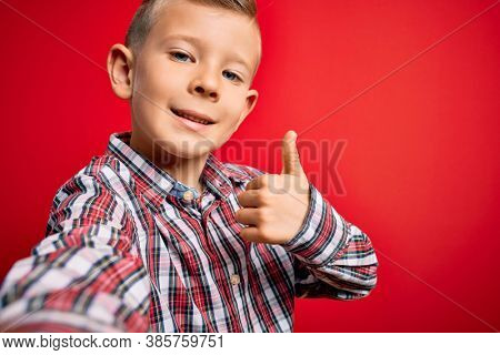Close up of young little caucasian kid with blue eyes taking a selfie photo over red background happy with big smile doing ok sign, thumb up with fingers, excellent sign