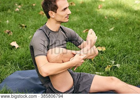 Young Athletic Man In Sportswear Doing Yoga In The Park. Practice Leg Cradle Asana Outdoors. People 