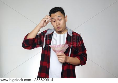 Confused Stressed Asian Man Holding Indonesian Rupiah Cash In Hand, Thinking Wondering How To Spend.