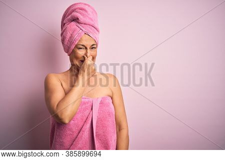 Middle age woman wearing bath towel from beauty body care over pink background smelling something stinky and disgusting, intolerable smell, holding breath with fingers on nose. Bad smell