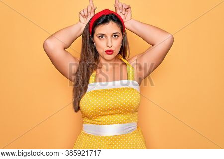 Young hispanic pin up woman wearing fashion sexy 50s style over yellow background doing funny gesture with finger over head as bull horns