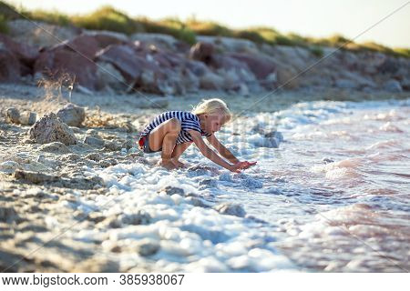 Little Girl Plays On The River Bank. She Touches The Water With Her Hands.