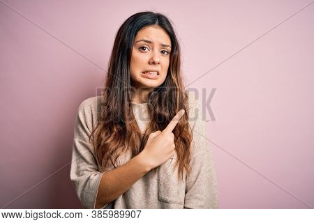 Young beautiful brunette woman wearing casual sweater standing over pink background Pointing aside worried and nervous with forefinger, concerned and surprised expression