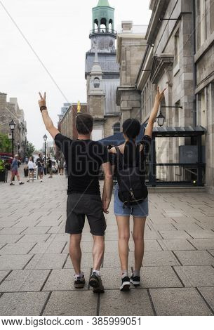 A Causasian Man And Chinese Woman Couple Flashing Peace Signs As They Walk Through Old Montreal In Q