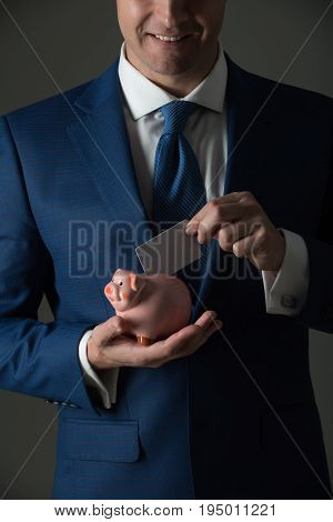 hands putting blank business card in piggy bank for saving on grey background. Stylish formal blue suit and tie. Banking money loan moneybox ecash and information