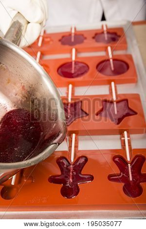 Sweets artisanal production of fruits loly pop at the factory. Close up of the saucepan pan with liquid hot red sugar dispensing to the orange mold. Artisanal production in France
